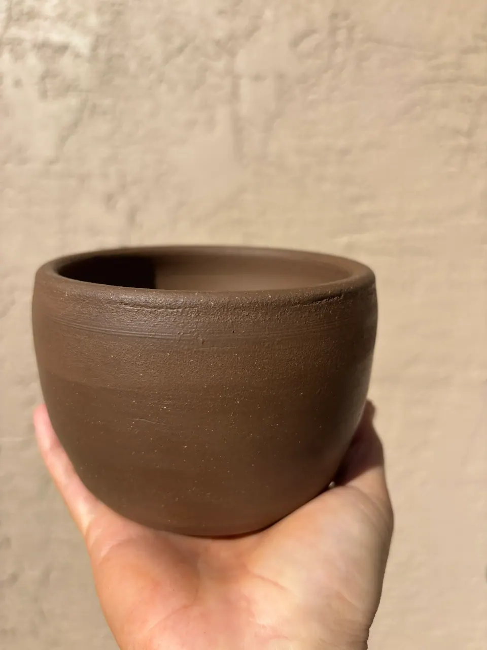 Unglazed dark brown clay bowl held in an open palm against a stucco wall