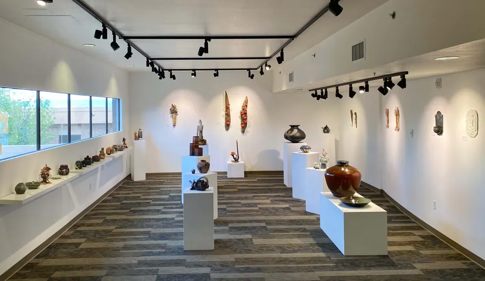 Gallery filled with ceramic vessels on white plinths and a long window shelf, with sculptural pieces hung on the walls