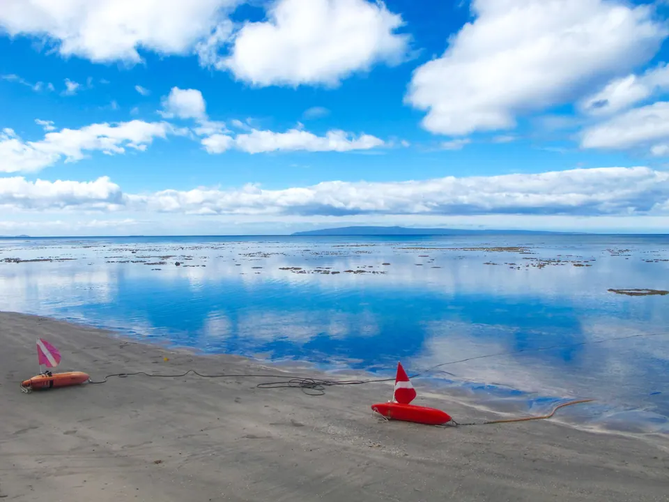 Glassy shoreline reflecting a blue sky full of clouds, with small red boats on the sand