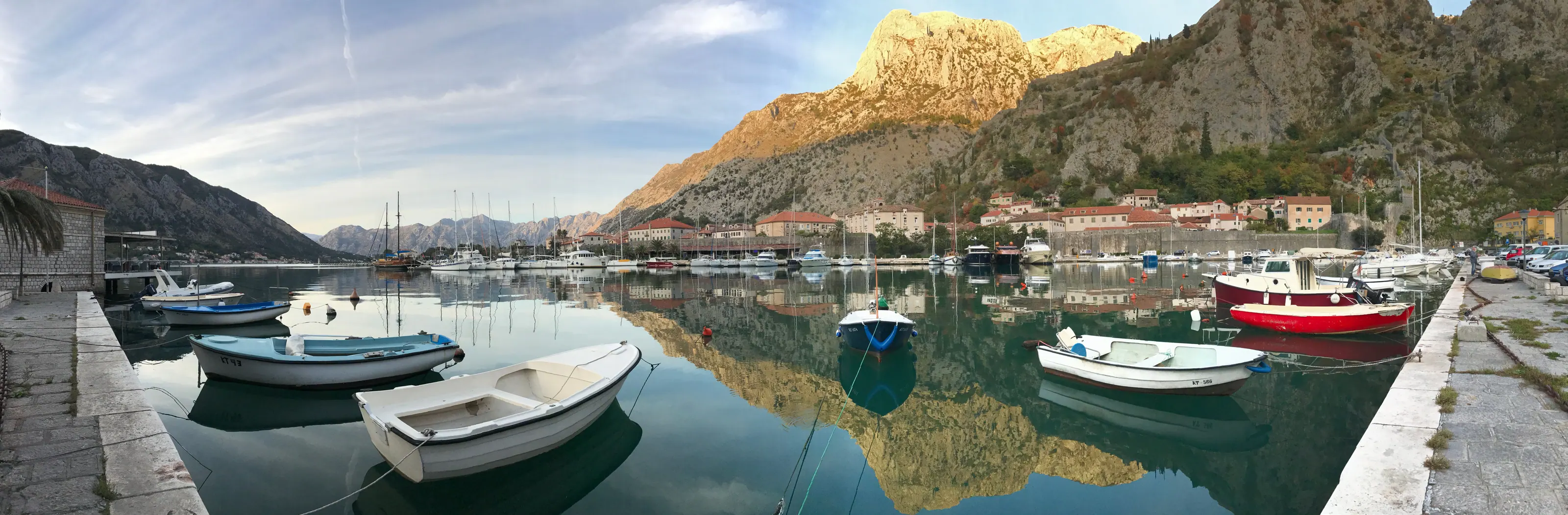 Wide panorama of a calm harbor with small moored boats beneath steep sunlit mountains
